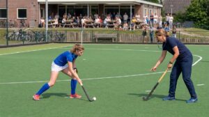 Een speelster oefent met de bal aan de stick, een trainer wijst de houding aan