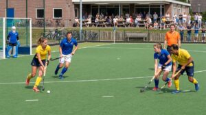 Vier spelers in een duel om de bal op het clubveld, de scheidsrechter kijkt toe
