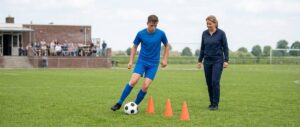Een speler oefent met de bal tussen de pionnen, een trainer kijkt toe