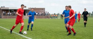 Vier spelers in een duel om de bal op het clubveld, de scheidsrechter kijkt toe